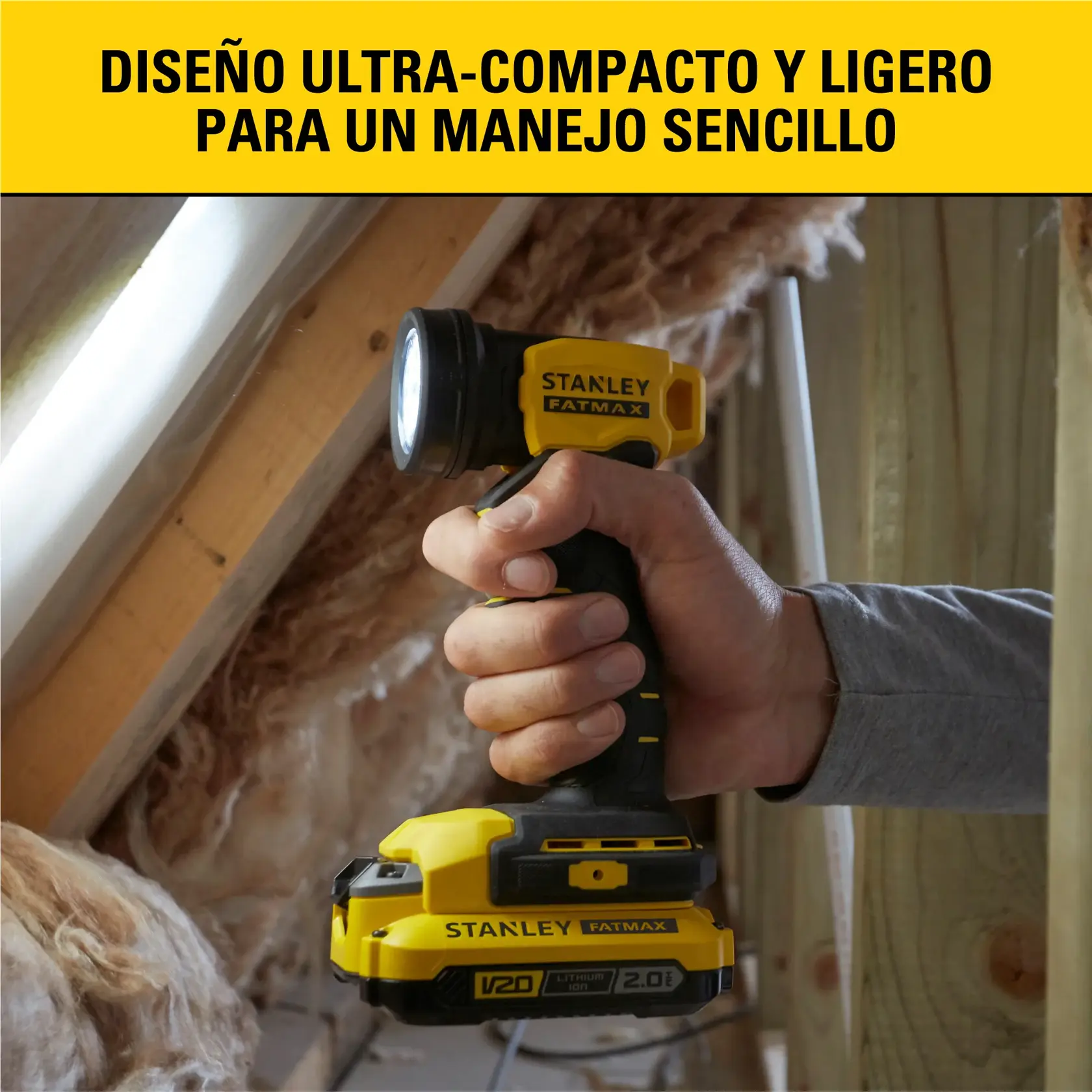 Una persona sostiene una linterna LED inalámbrica Stanley Fatmax V20 con un diseño compacto y ligero, adecuada para un manejo sencillo. El producto se muestra en un entorno de construcción o renovación, con aislamiento y vigas de madera al fondo.