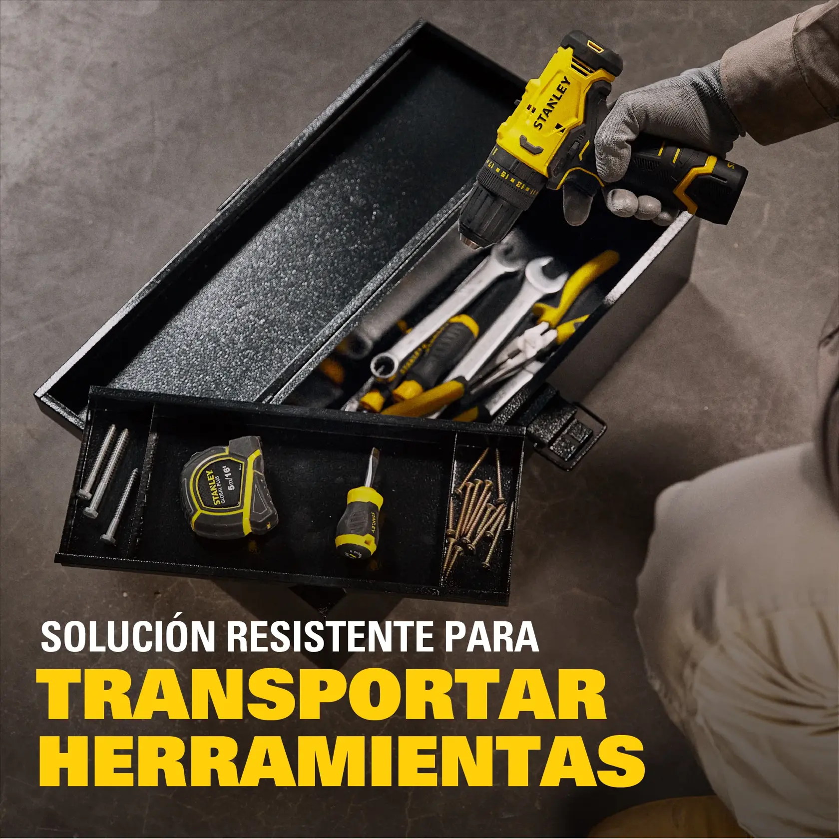 Se muestra una caja de herramientas negra de la marca STANLEY llena de herramientas manuales y tornillos variados. Una mano con guante sostiene un taladro inalámbrico amarillo y negro sobre la caja. Una cinta métrica, un destornillador y tornillos son visibles en la bandeja. El texto en español dice 'Solución resistente para transportar herramientas'.