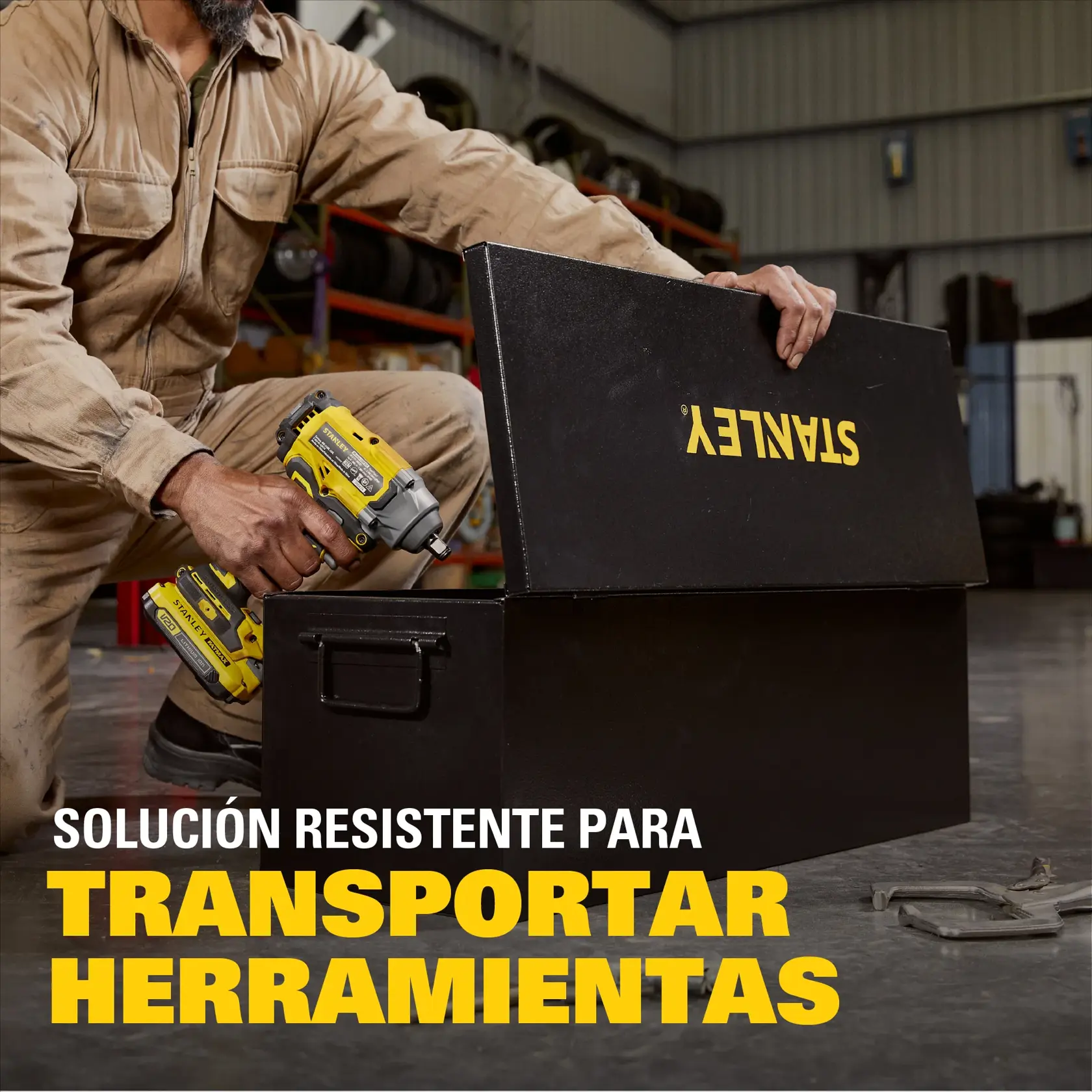 Una persona con mono beige de mecánico sostiene una herramienta eléctrica amarilla y abre una caja de herramientas negra STANLEY en un taller industrial. El texto en español dice: 'Solución resistente para transportar herramientas.'