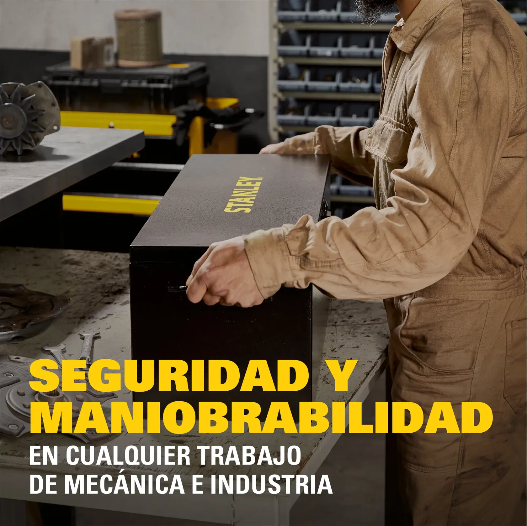 Una persona con traje de trabajo beige manipula una caja de herramientas negra con el logotipo amarillo de STANLEY sobre una mesa de trabajo en un taller industrial. El fondo muestra almacenaje de herramientas y estantes. El texto amarillo grande dice: 'Seguridad y maniobrabilidad en cualquier trabajo de mecánica e industria.'