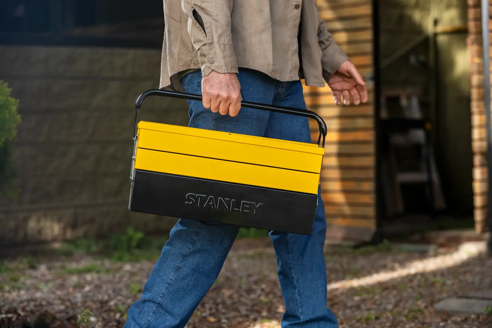 Caja de herramientas metálica STANLEY STMT98087-LA amarilla y negra llevada por una persona, vista lateral en exterior.