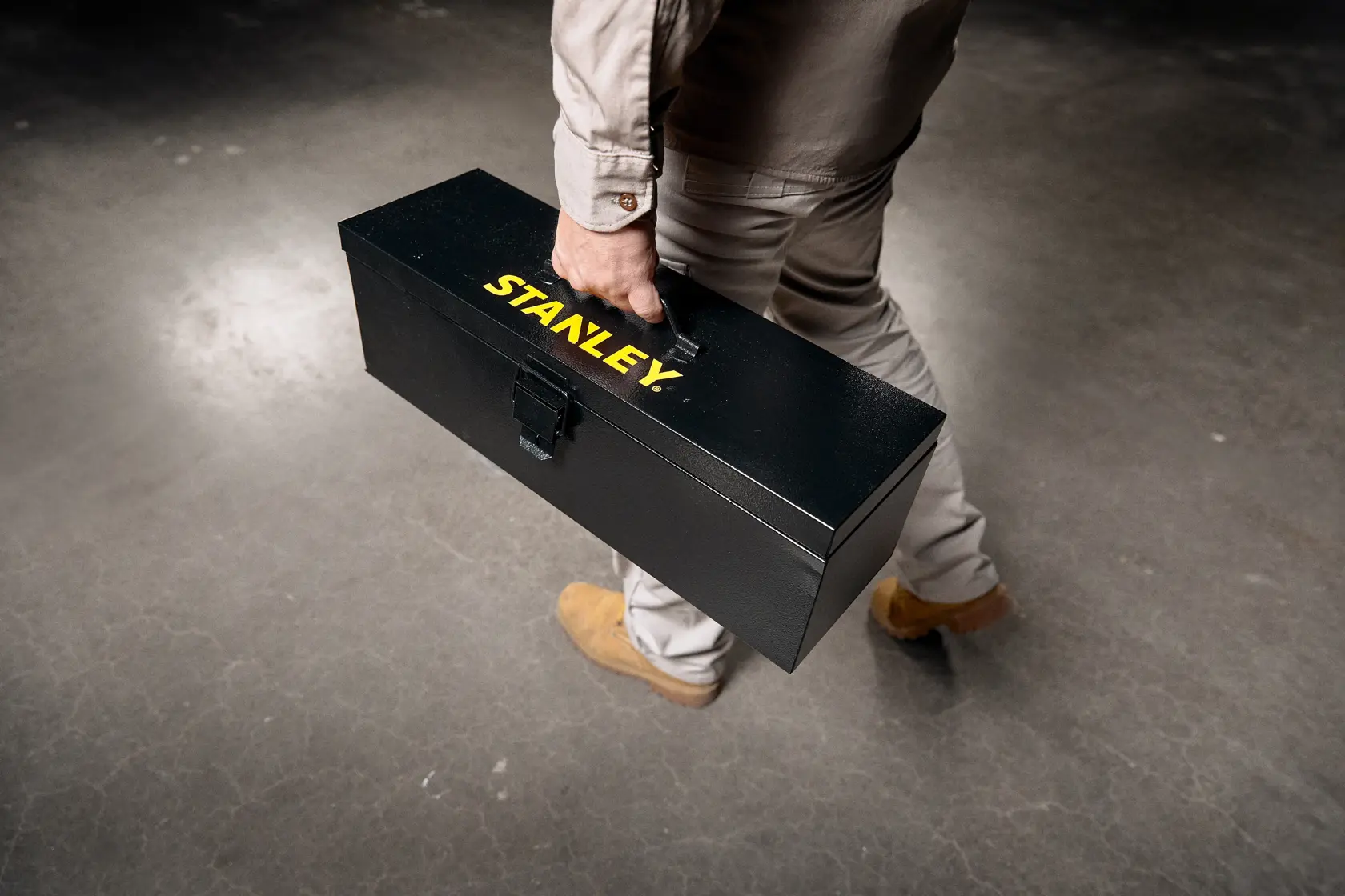 Stanley STST98726LA caja de herramientas metálica negra sostenida por una persona, fotografía de producto en piso de concreto.