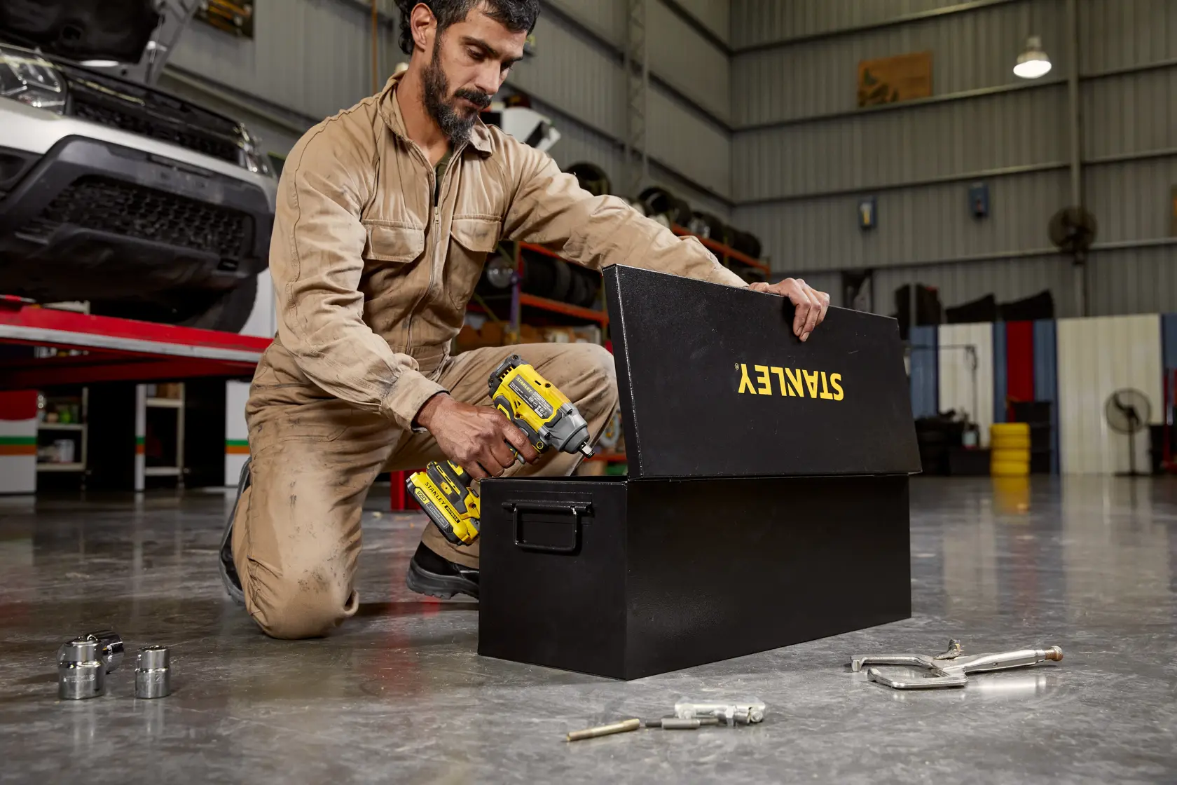 Caja de herramientas de metal negra STANLEY STST98727LA en un taller, mostrada abierta con herramientas y un trabajador al lado.